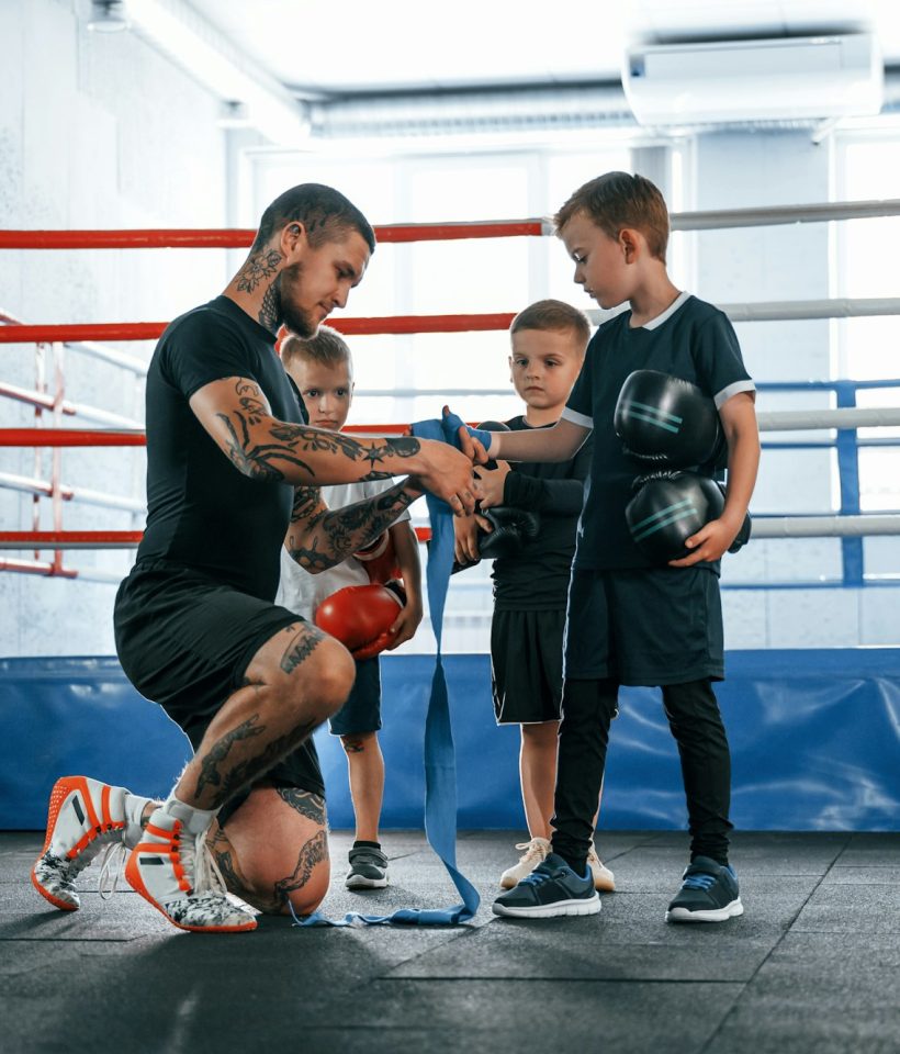 Wearing blue hand wraps. Young tattooed coach teaching the kids boxing techniques